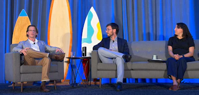 Three panelists seated on stage during a discussion with decorative surfboards on blue curtains backdrop