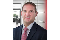Professional headshot of a smiling man in navy blazer and red patterned tie in modern office setting