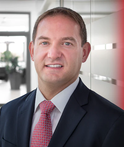 Professional headshot of a smiling man in navy blazer and red patterned tie in modern office setting