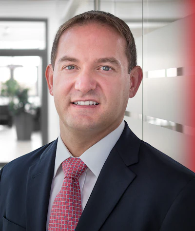 Professional headshot of a smiling man in navy blazer and red patterned tie in modern office setting